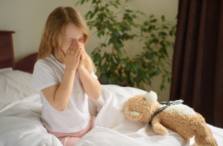 Young girl holding her nose on white bedding with teddy bear