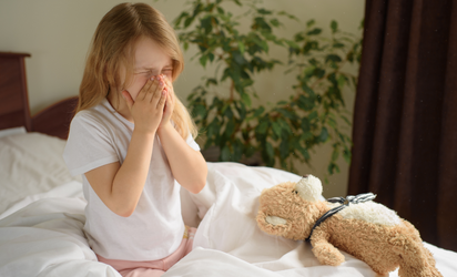 Young girl holding her nose on white bedding with teddy bear